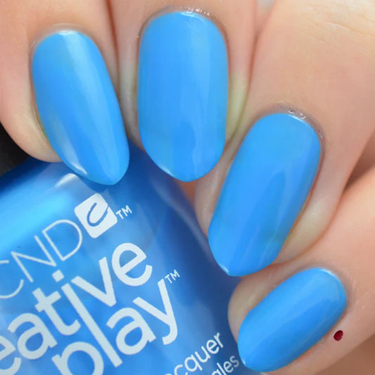 A close-up of fingernails painted in a bright medium blue shade, with part of the nail polish bottle visible in the foreground. The bottle label shows the brand name CND and the line name Creative Play.