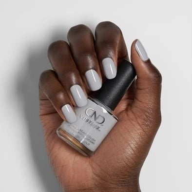 A hand with medium-dark skin holding a bottle of CND Vinylux #184 Thistle Thicket nail polish. The nails are painted a solid, glossy light gray.