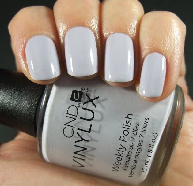 A close-up of a hand holding a bottle of CND Vinylux #184 Thistle Thicket showing the nails painted in a glossy pale lavender-gray shade.