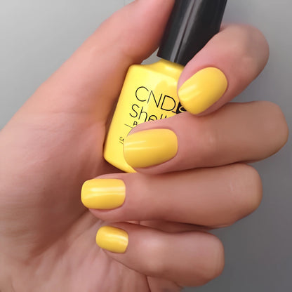 A hand with neatly manicured yellow nails holding a bottle of CND Shellac Bicycle yellow nail polish against a plain gray background.