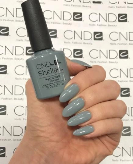 A hand holding a bottle of CND Shellac Gel Polish Mystic Slate nail polish in a dusty light gray-blue shade, with the fingernails painted to match, against a white background with a repeating CND logo.