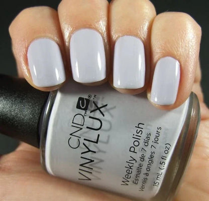 A close-up of a hand holding a bottle of CND Vinylux #184 Thistle Thicket showing the nails painted in a glossy pale lavender-gray shade.