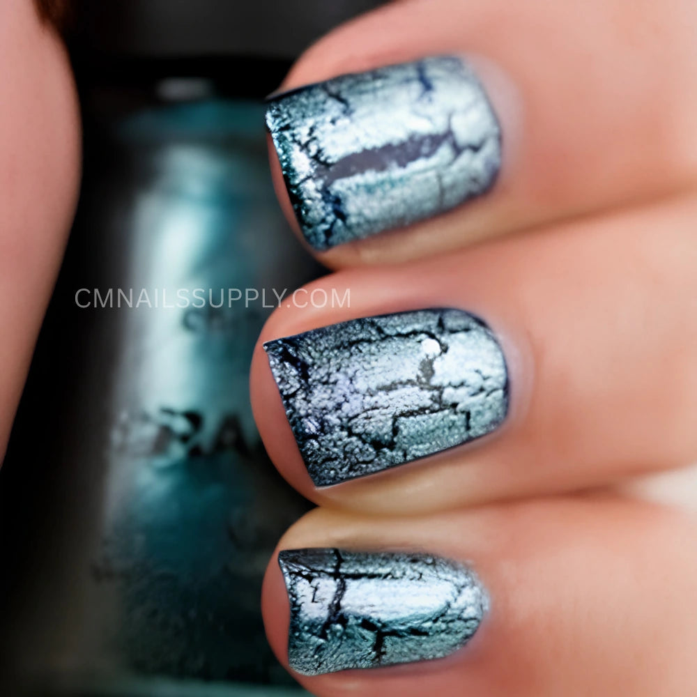 Close-up of a hand with fingernails painted in a metallic teal-blue shade with a black crackle pattern, with a matching nail polish bottle blurred in the background. A watermark reads CMNAILSSUPPLY. China Glaze Crackle Nail Polish #1047 Oxidized Aqua