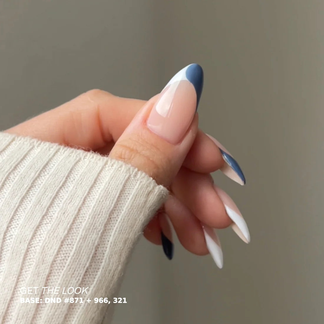 A close-up of a hand with long almond-shaped nails styled in a French tip manicure with a navy blue accent, wearing a cream ribbed knit sweater against a neutral gray background.