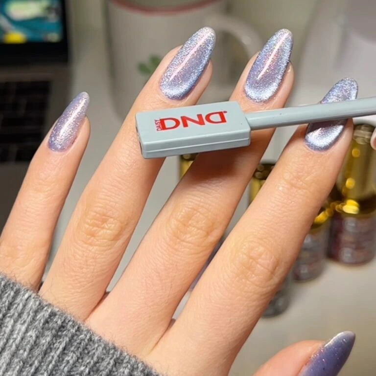 A close-up of a hand with long, manicured nails painted in a shimmering lavender-purple cat-eye polish, holding a gray cat eye magnet.