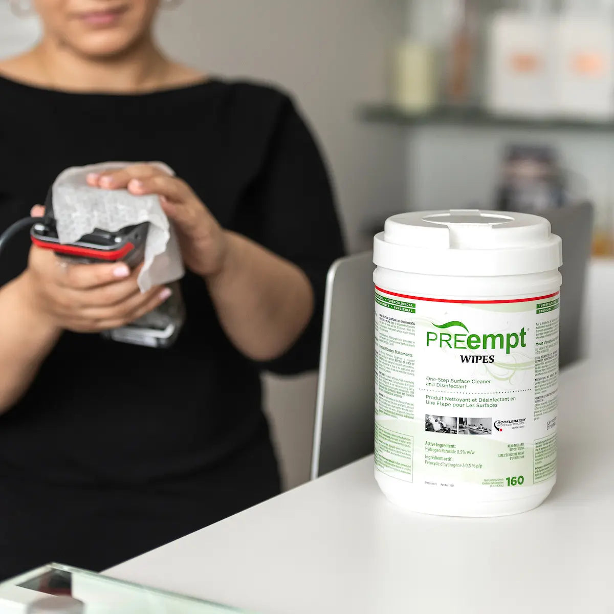 Person using a handheld device with a container of Preempt Wipes on a desk.