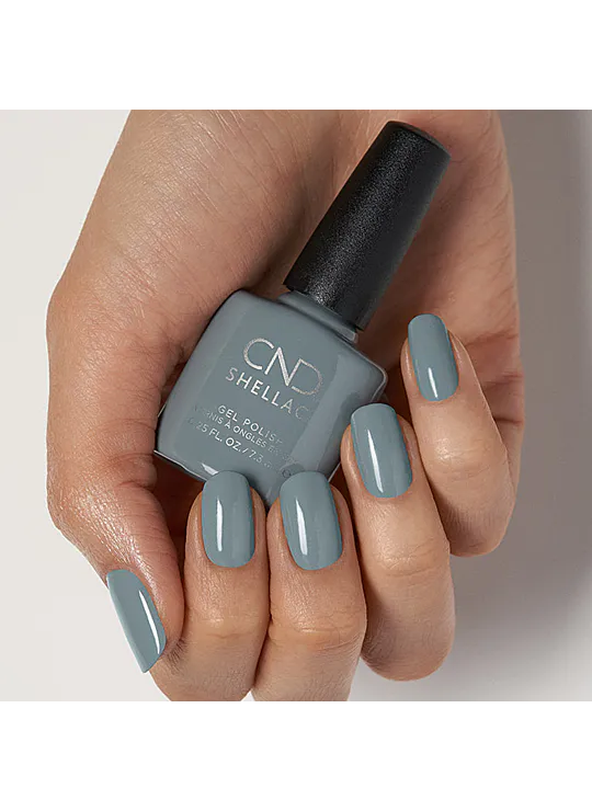 A hand holding a bottle of CND Shellac gel nail polish Mystic Slate in a muted slate blue-gray shade, with the nails painted to match.