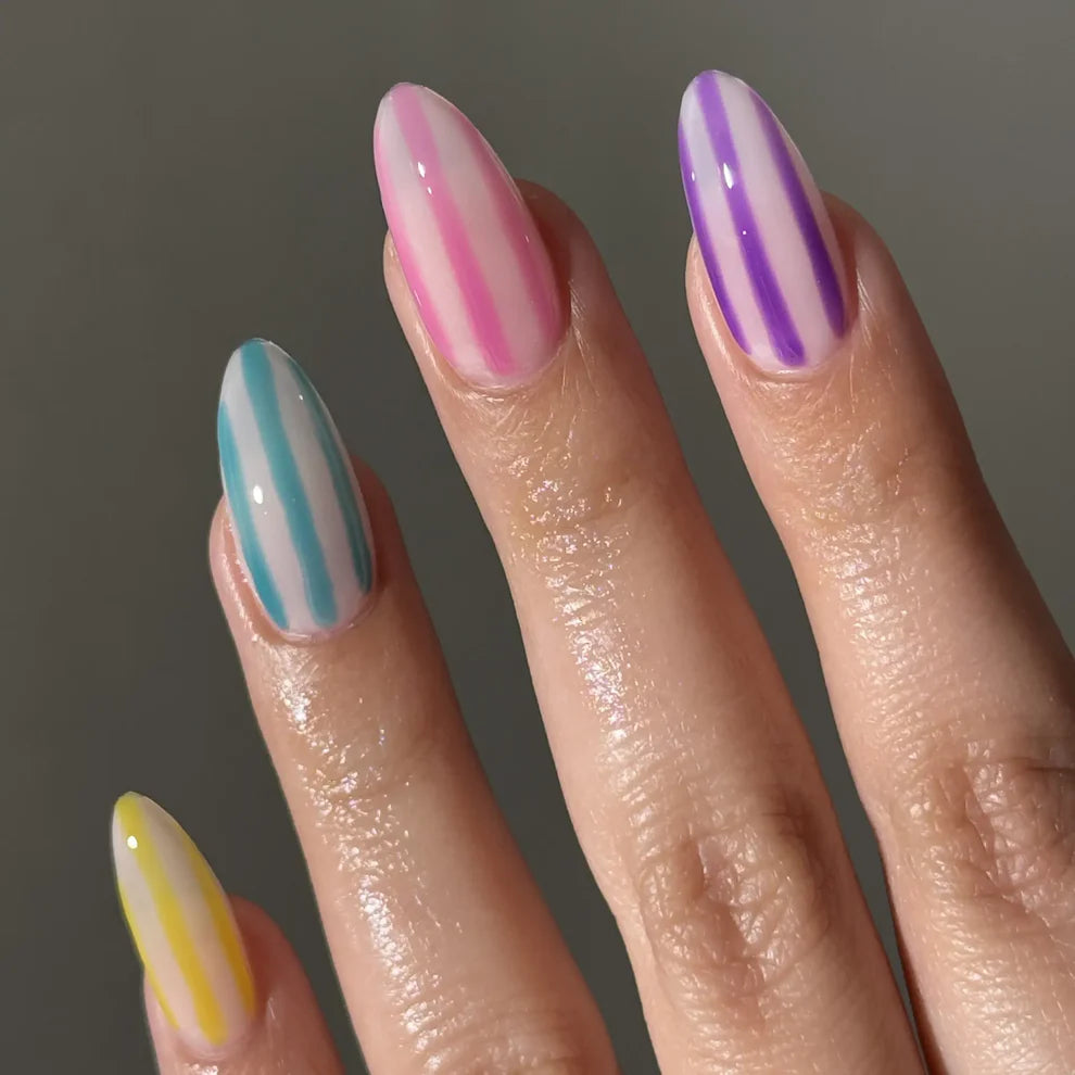 A close-up of a hand showcasing manicured almond-shaped nails with pastel vertical stripes. The nails feature pink, purple, blue, and yellow stripes on a neutral gray background, with DC Jelly #03 Yuzu manicure featuring a lemon yellow jelly nail polish finish.