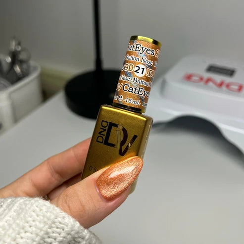 Hand with a caramel orange cat eye manicure holding the DND Diva Cat Eye Gel Polish #21 Button Nose bottle.