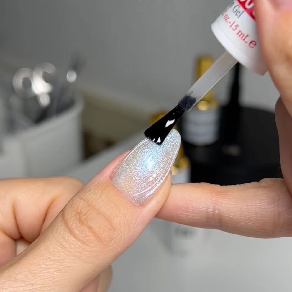 A close-up of a DND gel top coat polish being applied to a fingernail for manicure. The nail polish is a shimmering, iridescent light silver with a subtle holographic finish, and the brush is being held by a hand while the bottle is held by another hand.