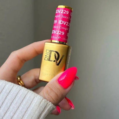 Gold nail polish bottle with pink cap held by a hand with bright pink nails against a neutral background