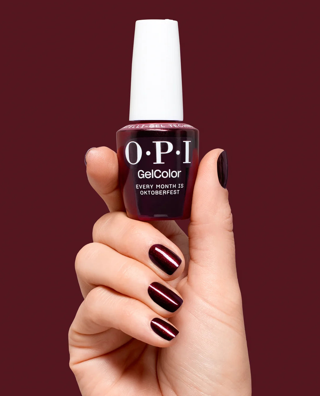 Hand holding a bottle of OPI GelColor Every Month is Oktoberfest gel nail polish with a dark red color on a matching background