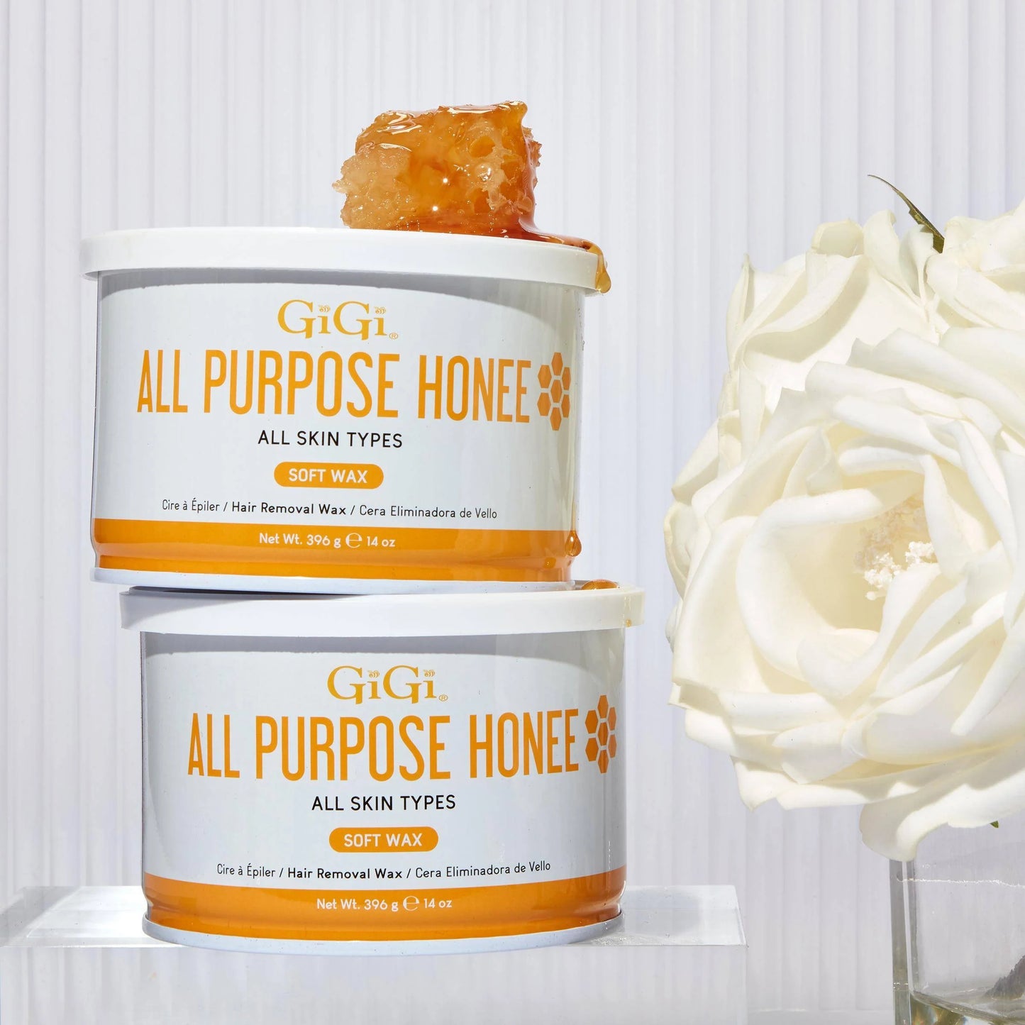 Two tubs of GiGi All Purpose Honey soft wax stacked on a white surface, with a piece of honey on top of the upper tub and a large white rose beside them against a white paneled background.
