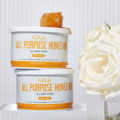 Two tubs of GiGi All Purpose Honey soft wax stacked on a white surface, with a piece of honey on top of the upper tub and a large white rose beside them against a white paneled background.