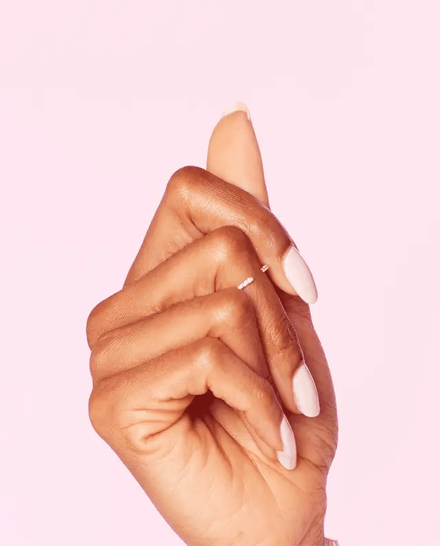 A close-up of a hand with long, almond-shaped pink OPI Let me Bayou a Drink manicured nails, wearing a delicate silver ring on one finger, set against a solid light pink background.