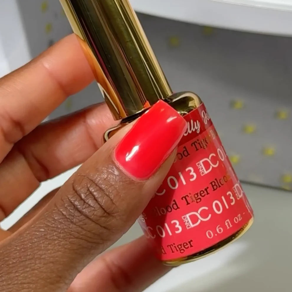 Manicured hand holding the DC Jelly 013 Tiger Blood jelly nail polish bottle, showcasing the fiery orange red color on the nails