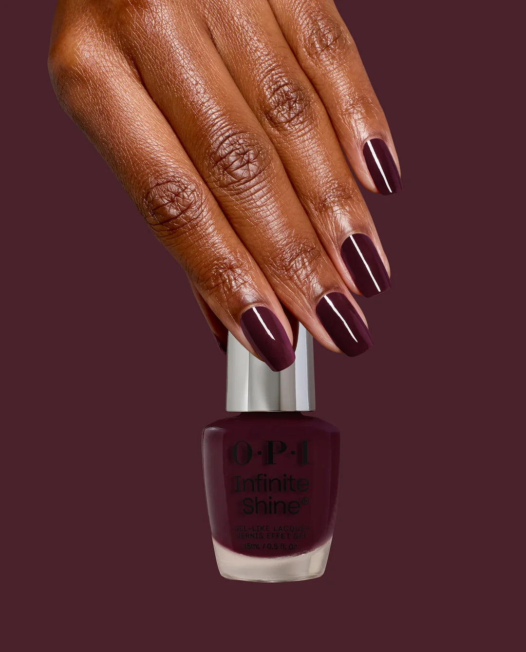 A hand with glossy dark plum nail polish holding an OPI Infinite Shine Mrs. O’ Leary’s BBQ bottle of the same shade against a solid dark plum background.